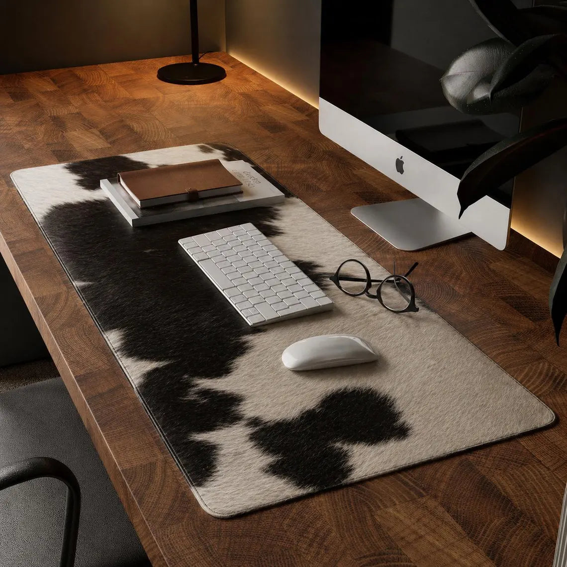A large rectangular desk mat with a black and cream cowhide pattern is styled on a wooden desk, surrounded by a keyboard, mouse, glasses, and a computer monitor.