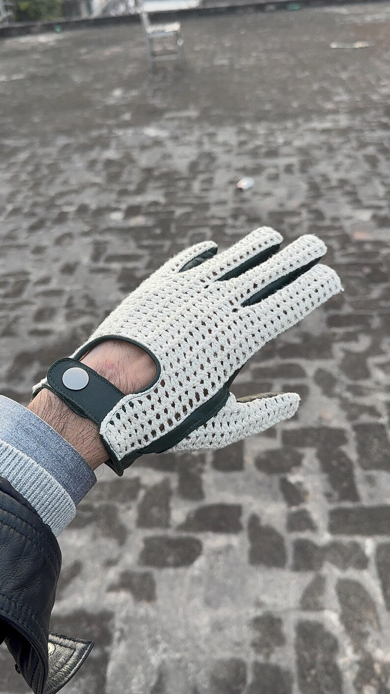 A white knit golf glove with black trim and a black wrist strap featuring a silver snap button, worn on a person's hand against a blurred cobblestone background.