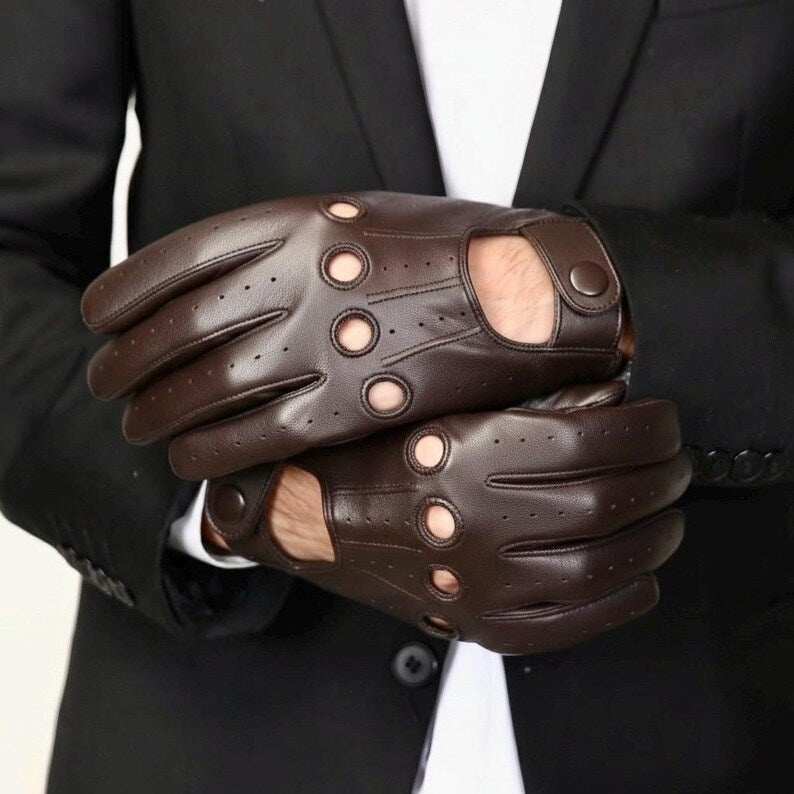 A pair of dark brown leather semi-open gloves with circular cutouts, worn on a person's hands in front of a black suit jacket and white shirt.