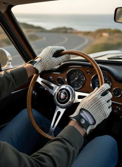 A person driving a vintage car, wearing light beige knit perforated gloves with black fingertips. The interior of the car features a wood-grain dashboard and a classic three-spoke steering wheel with a visible red and white logo.