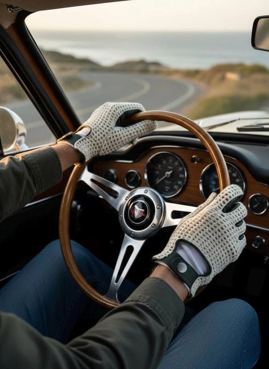 A person driving a vintage car, wearing light beige knit perforated gloves with black fingertips. The interior of the car features a wood-grain dashboard and a classic three-spoke steering wheel with a visible red and white logo.