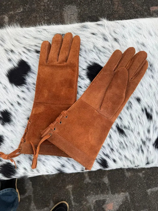 A pair of long brown suede gloves laid flat on a black-and-white cowhide rug, with a dark paved surface visible around the edges.