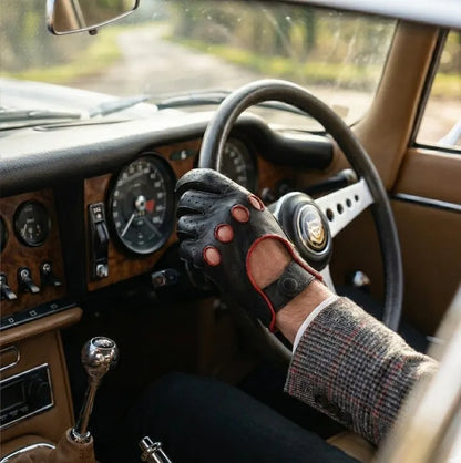 A close-up of a person's hand wearing a black leather driving glove with red trim and finger cutouts, resting on the steering wheel inside a vintage car cabin.