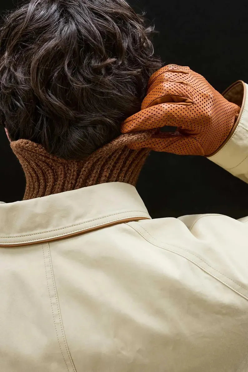 A close-up lifestyle shot of a person from behind, wearing a cream-colored leather jacket, a brown ribbed knit neck warmer, and textured brown leather gloves while adjusting the neck warmer.