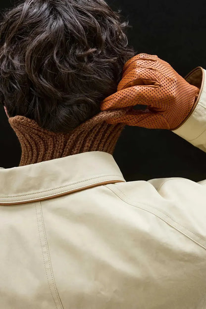 A close-up lifestyle shot of a person from behind, wearing a cream-colored leather jacket, a brown ribbed knit neck warmer, and textured brown leather gloves while adjusting the neck warmer.
