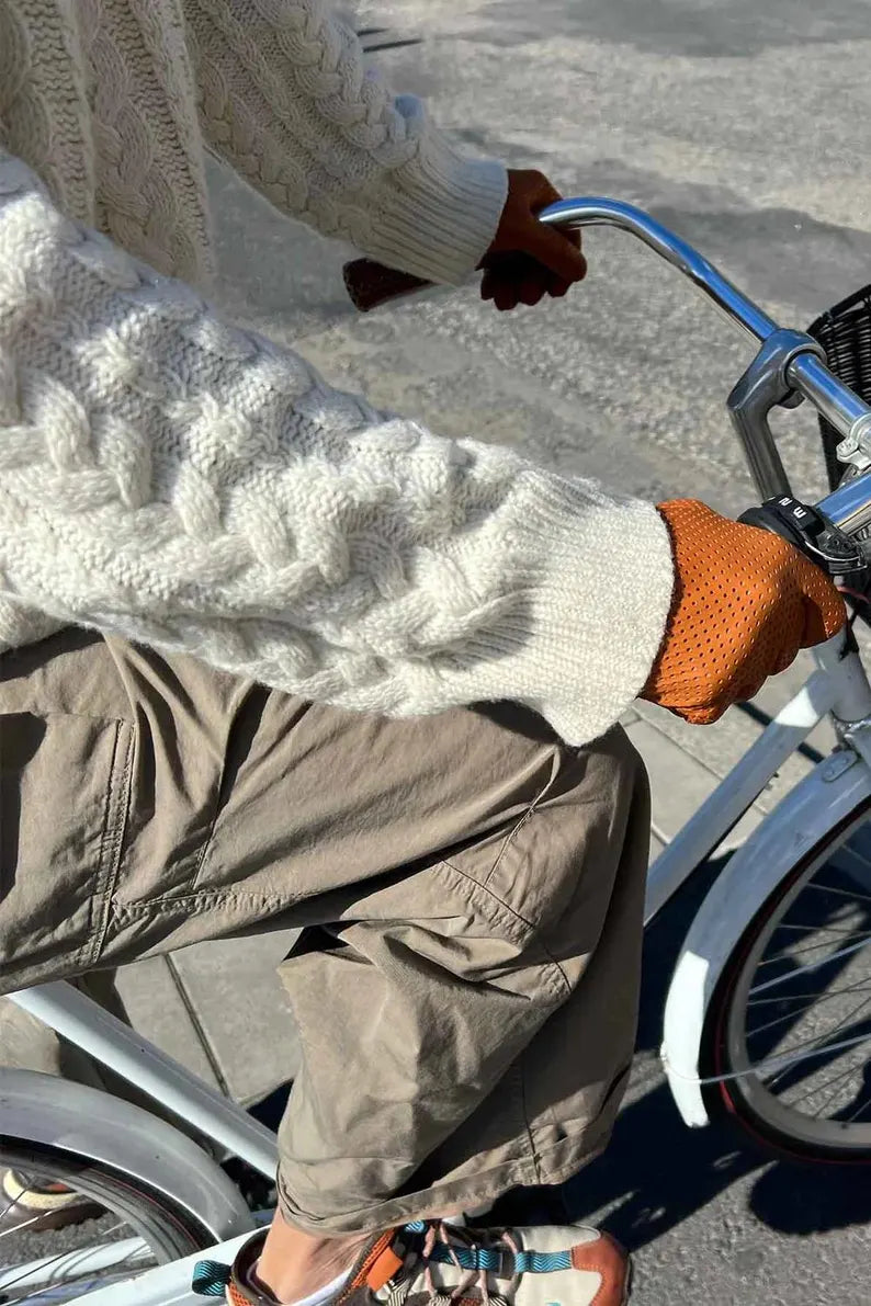 A person riding a bicycle outdoors, wearing a cream cable-knit sweater, brown cargo pants, and orange knit gloves.