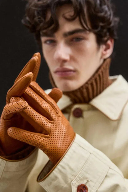 A close-up of a man wearing a cream-colored jacket and a brown ribbed turtleneck sweater, holding up a pair of brown leather gloves with perforated detailing.