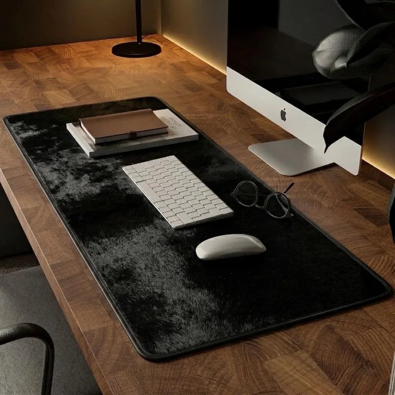 A large black desk mat with a textured, plush-like surface is laid out on a wooden desk. The mat is styled with a white keyboard, a white mouse, a stack of notebooks, and a pair of eyeglasses.
