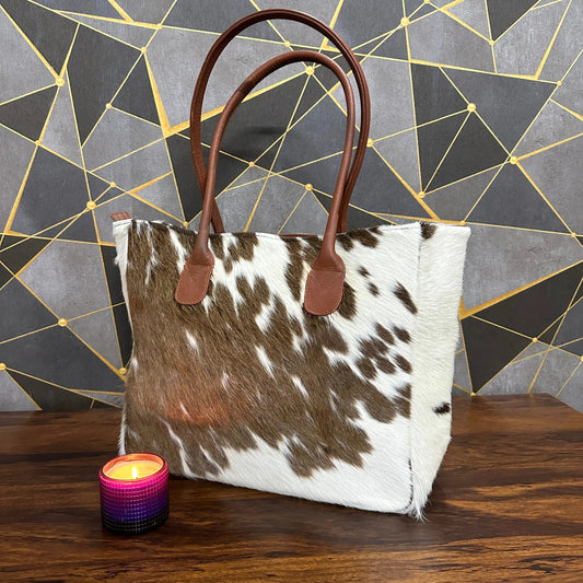 Tote bag with cowhide pattern and brown handles on a wooden surface with a geometric-patterned wall in the background.