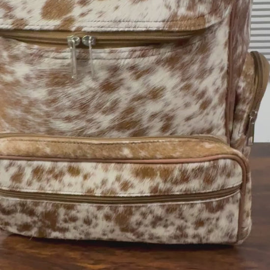 360 veiw of A brown and white cowhide-patterned backpack with matching faux-leather trim, shown standing on a dark wooden table in front of a window with horizontal blinds.