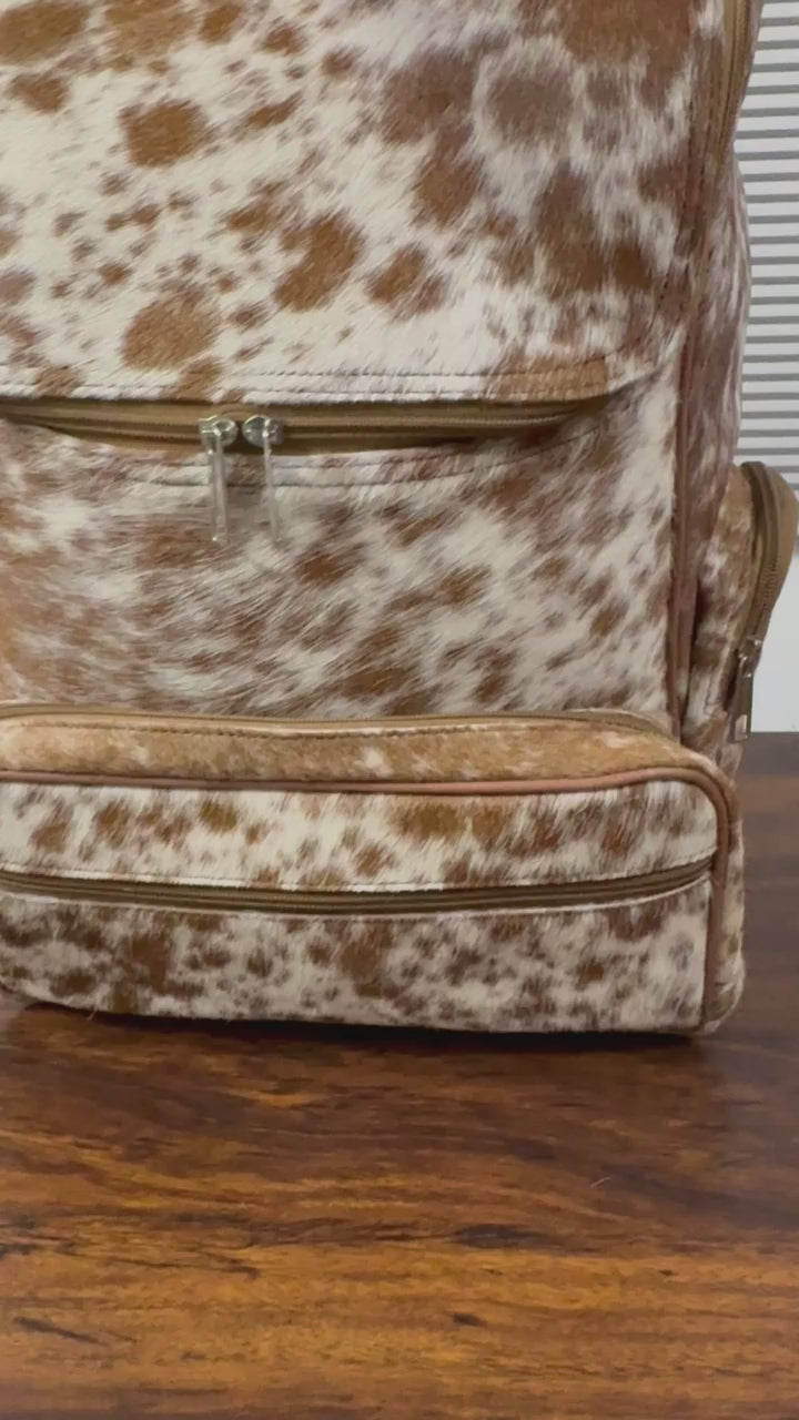 360 veiw of A brown and white cowhide-patterned backpack with matching faux-leather trim, shown standing on a dark wooden table in front of a window with horizontal blinds.