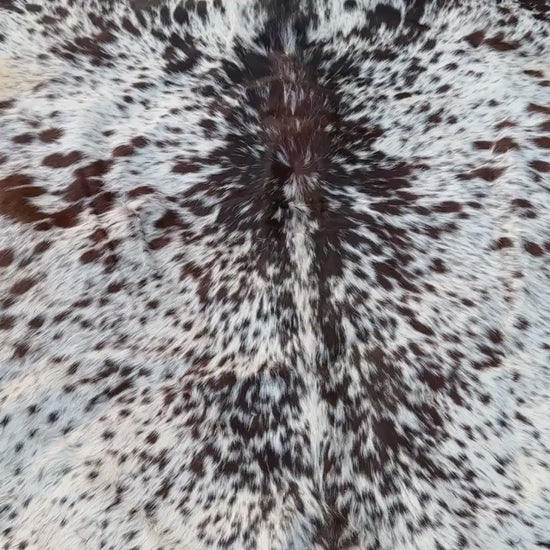 A natural cowhide rug with a black and white spotted pattern and tan edges, laid flat on a green grass lawn.
