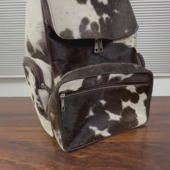 360 veiw of A cowhide backpack with a black and white spotted pattern, shown standing on a wooden table in front of a window with horizontal blinds.