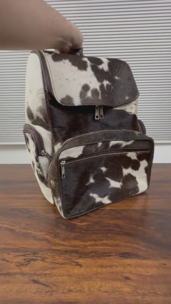 360 veiw of A cowhide backpack with a black and white spotted pattern, shown standing on a wooden table in front of a window with horizontal blinds.